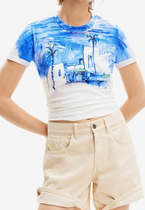 Persona con una camiseta blanca y azul con un dibujo de una palmera y un edificio, combinada con pantalones cortos beige remangados.