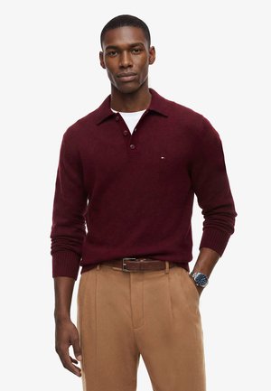 Man draagt een bordeauxrode lange-mouwen polotrui over een wit ondershirt, beige broek, bruine riem en een zilveren horloge met blauwe wijzerplaat.