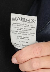 Pesulappu kankaalla, jossa on symboleja pesu-, kuivaus- ja silitysohjeista. Vaaleanharmaa tausta mustalla tekstillä. Materiaali on kangasta.