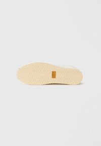 Semelle de chaussure avec motif de bande de roulement beige en zigzag et petit patch logo rectangulaire marron au centre, sur fond clair uni.