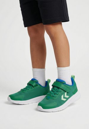 Barn iført grønne og blå sneakers med hvide såler, hvide sokker og sorte shorts, stående på en ensfarvet hvid baggrund.