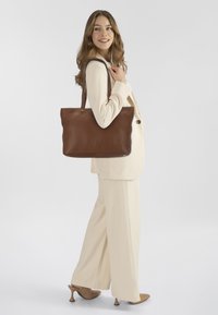 Bruine leren tote bag met een gestructureerde afwerking, twee schouderbanden en een gladde ontwerp. Model draagt een crèmekleurig pak en hakken.