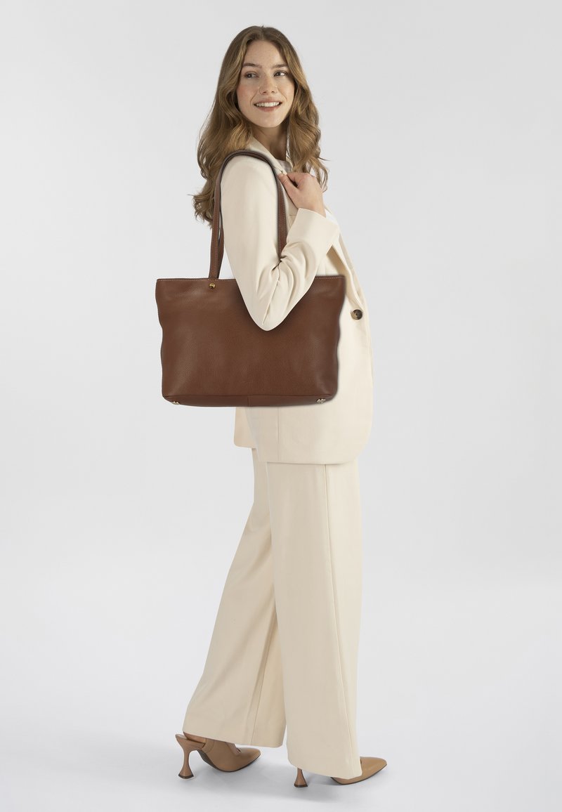 Bruine leren tote bag met een gestructureerde afwerking, twee schouderbanden en een gladde ontwerp. Model draagt een crèmekleurig pak en hakken.