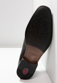 Suela de zapato negra con goma texturizada, con un inserto de cuero marrón y un logo grabado. Detalles de texto grabados incluidos en el reverso.