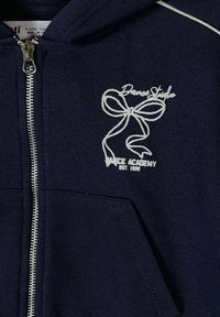 Sweat à capuche zippé bleu marine avec un nœud blanc brodé et le texte "Dance Studio Dance Academy Est. 1978" sur la zone de la poche poitrine gauche.