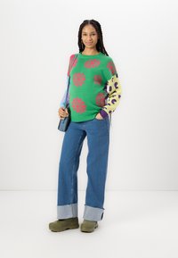 Πράσινο πουλόβερ με ροζ λουλουδάτα σχέδια, πολύχρωμα μανίκια, σε συνδυασμό με ανοιχτόχρωμα μπλε τζιν και ελαιούχια πράσινα παπούτσια με πλατφόρμα.