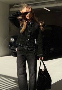 Veste en denim noir avec des boutons blancs, associée à un jean noir à jambes larges. Un sac fourre-tout noir est tenu dans une main, avec un design simple.