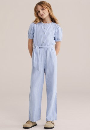 Fată tânără cu păr blond lung, purtând o salopetă albastru deschis cu dungi, nasturi și cordon în talie, stând cu mâinile la spate.