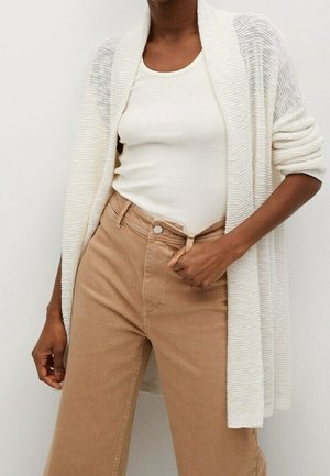 Personne portant un débardeur blanc côtelé, un cardigan tricot beige clair et un pantalon taille haute couleur camel, ajustant la taille d'une main.