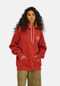 Červená mikina s kapucňou a sťahovacími šnúrkami, predné vrecko a grafický logo na hrudi, kombinovaná s nohavicami s leopardím vzorom. Mäkkučká tkanina, voľný strih.