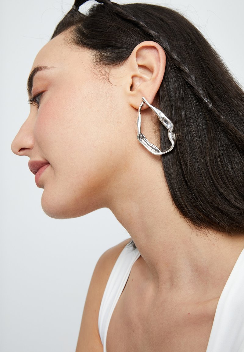 Grands créoles argentées avec un design ondulé, présentant une finition polie et des accents texturés, portées à l'oreille d'un mannequin.