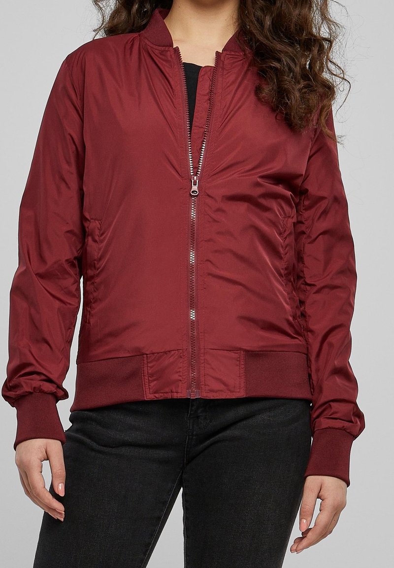 Personne portant une veste zippée rouge foncé avec poignets et ourlet côtelés, associée à un pantalon noir sur un fond uni.