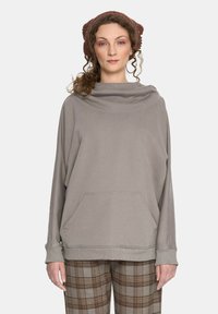 Jasnoszara oversize bluza z kapturem, długimi rękawami, bocznymi kieszeniami i ściągaczami, noszona z brązowymi spodniami w kratę.