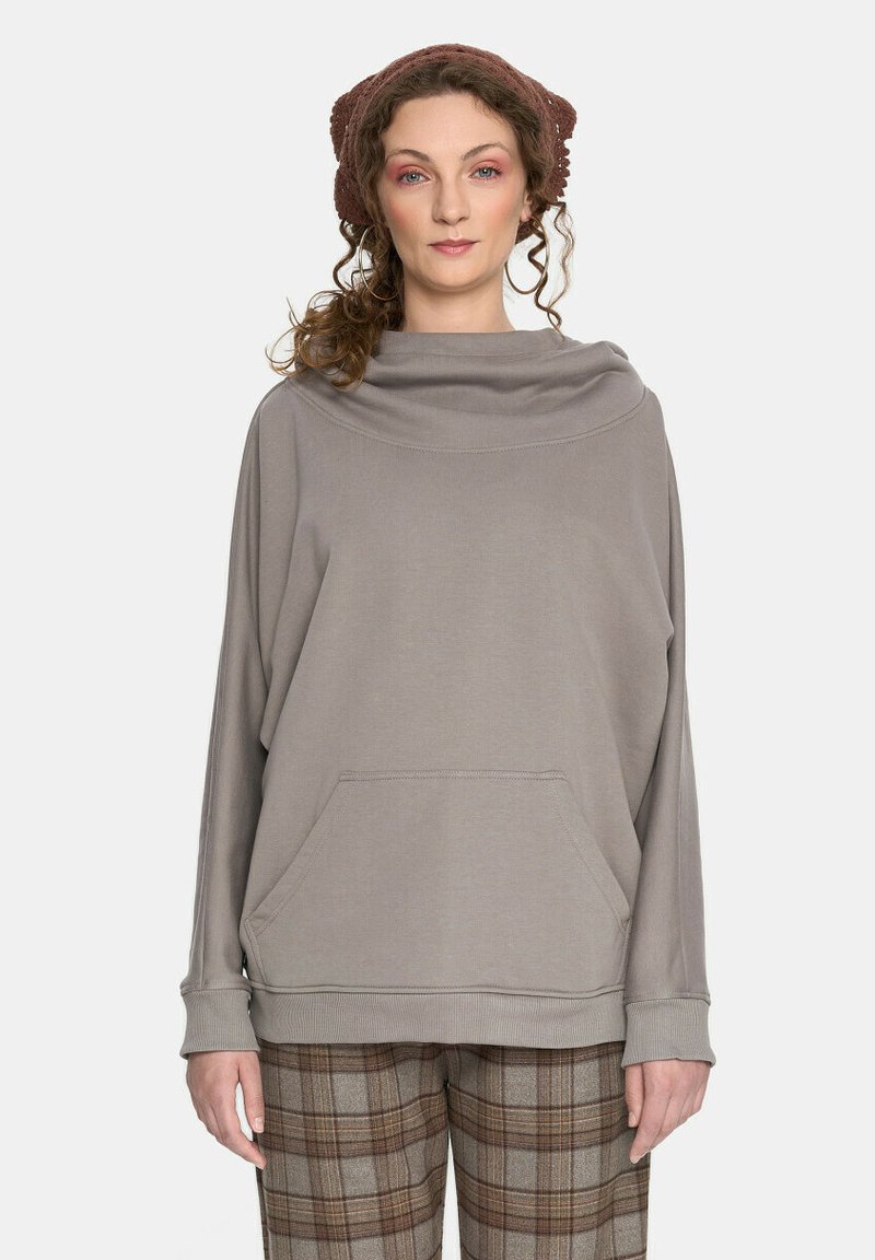 Jasnoszara oversize bluza z kapturem, długimi rękawami, bocznymi kieszeniami i ściągaczami, noszona z brązowymi spodniami w kratę.