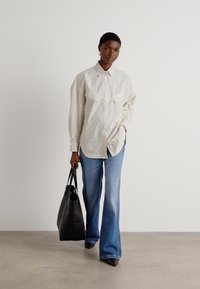Camicia oversize a strisce in colori chiari, abbinata a jeans svasati blu chiaro. Il modello tiene una borsa a tracolla nera. Tessuto dalla texture liscia.