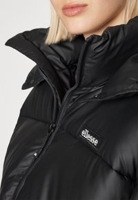 Czarna kurtka puchowa z wysokim kołnierzem, wyposażona w przedni suwak i wyraźne logo ellesse. Gładka tekstura oraz widoczny pikowany design.