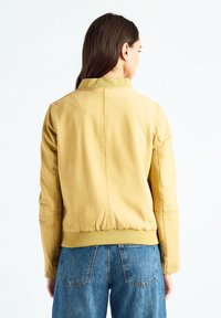 Veste en cuir jaune avec col rond, poignets côtelés et taille élastique. Texture lisse avec des détails de couture minimaux.