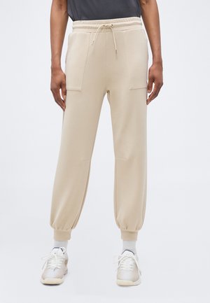 Osoba ubrana w beżowe spodnie dresowe z elastycznymi mankietami, dużymi przednimi kieszeniami oraz białe tenisówki na jednolitym tle.