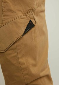 Tissu de pantalon cargo beige avec une texture lisse. Dispose d'une poche latérale avec un écusson triangulaire noir et des coutures contrastantes orange.