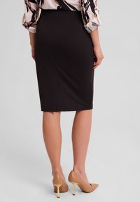 Czarna spódnica ołówkowa o gładkiej fakturze, dopasowana sylwetka, subtelne rozcięcie z tyłu, zestawiona z nude szpilkami i wzorzystą bluzką.