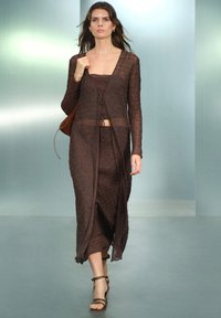 Braun gestricktes Kleid mit langen Ärmeln, V-Ausschnitt und vorderem Bindedetail. Transparente Textur, bodenlang, kombiniert mit braunen Sandalen mit Riemen und einer braunen Tasche.