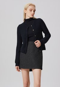 Cardigan en tricot bleu marine avec un tissu texturé, col rond, boutons sur le devant, et une forme ajustée associé à une jupe à motifs.