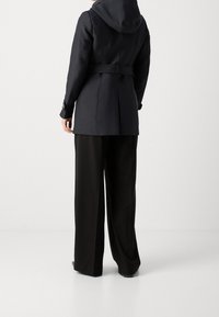 Veste imperméable bleu marine avec capuche, dotée d'une taille ceinturée, d'une ventilation au dos et de poignets à double bouton, associée à un pantalon noir ample.