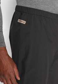 Pantalons de sport noirs avec une taille élastique, comportant une petite étiquette de logo sur la poche arrière. Le matériau semble lisse et léger.