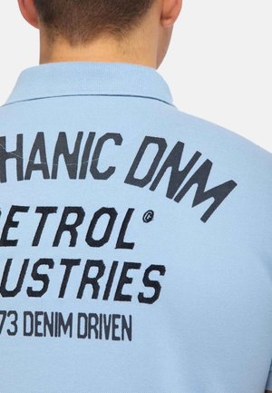 Personne portant un polo bleu clair avec un texte noir dans le dos portant la mention "MECHANIC DNA PETROL INDUSTRIES 73 DENIM DRIVEN."