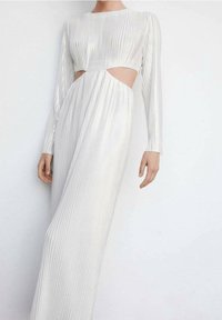 Robe blanche à manches longues avec des plis verticaux, présentant des découpes sur les côtés. Le tissu a une texture lisse et brillante.
