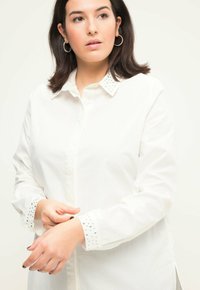 Hvid skjorte med oppe på det udsmykkede krave og manchetter med sølvnitter, lange ærmer og en lige skæring.