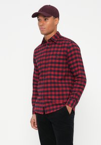 Man met een rood en zwart geruit overhemd met knopen, zwarte broek en een donkerrode baseballcap, staande met één hand in zijn zak.