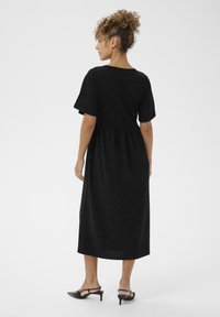 Vestido negro de media longitud con mangas cortas abiertas, cintura ajustada y falda fluida. El material tiene una textura suave. Se lleva con zapatos de tacón negros.