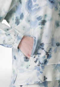 Sweatshirt cu model floral în bleu deschis și alb, cu o textură moale, manșete elasticizate și un buzunar frontal pentru funcționalitate.