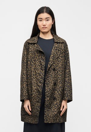 Femeie cu păr lung și închis la culoare, purtând o haină de leopard maro până la genunchi peste o rochie neagră, pe un fundal alb simplu.