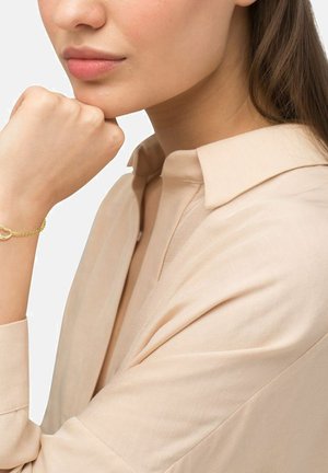 Mujer de tono de piel clara reposando el mentón sobre el puño, vistiendo una camisa beige con cuello y una delicada pulsera de cadena de oro con un encantos.