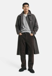Cappotto houndstooth marrone con ampio colletto, maglione grigio e pantaloni scamosciati marroni. Una cintura nera e scarpe scure completano l'outfit.