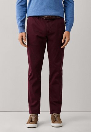 Hombre con pantalones burdeos, jersey azul, cinturón trenzado marrón y zapatillas de ante marrón de pie frente a un fondo liso.