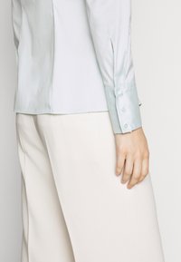 Blusa di raso azzurro chiaro con una texture liscia, maniche lunghe e polsini con bottoni, abbinata a pantaloni color crema.