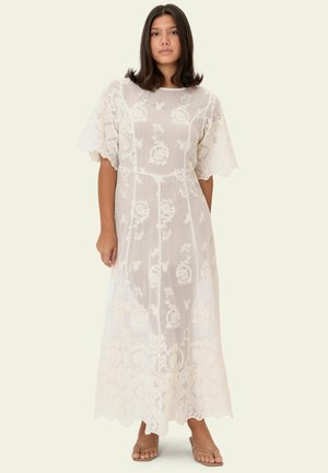 Fehér hímzett maxiruha áttetsző anyaggal, virágmintákkal, kerek nyakkivágással és fodros ujjakkal, bézs szandálokkal párosítva.