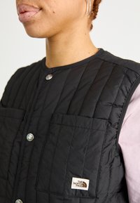 Chaleco negro acolchado con cuello redondo, botones a presión metálicos y dos bolsillos frontales; presenta un pequeño logo de la marca en la esquina inferior derecha.