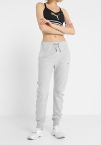 Schwarzer Sport-BH mit goldenem Logo und weißen Streifen, kombiniert mit grauen Jogginghosen mit Kordelzug und weißen seitlichen Akzenten. Weiße Schuhe.