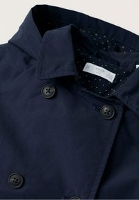 Mörkblå trenchcoat med klassisk krage, dubbelknäppt design och svarta knappar. Fodret har ett mönster med prickar.