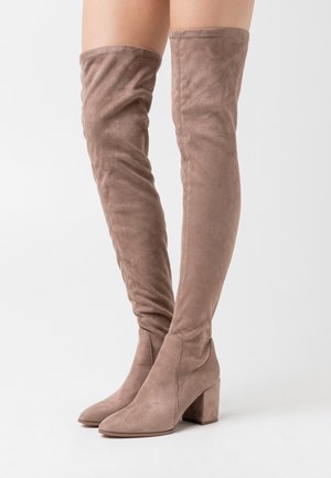 Bottes en daim taupe montant au-dessus du genou avec talons carrés, portées sur des jambes nues contre un fond clair uni.