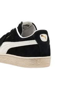 Sneaker Puma in suede nero con striscia bianca, suola texturizzata beige e logo Puma dorato sul tallone e sulla linguetta.