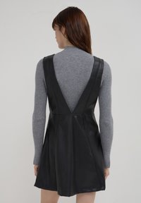 Schwarzes Lederkleid mit V-Ausschnitt und breiten Trägern, das einen figurbetonten Oberteil und einen ausgestellten Rock hat, getragen über einem grauen, gerippten Pullover.