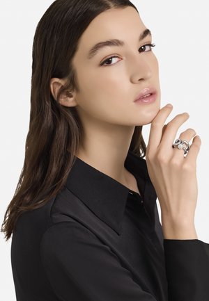 Srebrn prstan z teksturiranim dizajnom, ki ima osrednji dragulj, nosi se na roki z lepo urejenimi nohti. Model nosi črno srajco s ovratnikom.