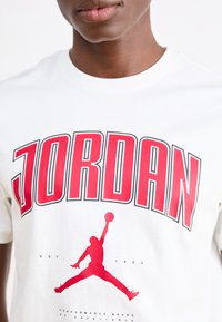 Biela bavlnená tričko s výrazným červeným textom "JORDAN" a čiernou siluetou skákajúcej postavy držacej basketbal pod ním.