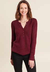 Maglione lavorato a maglia color bordeaux con scollatura a V, texture a coste e patta con cinque bottoni. Design aderente con maniche lunghe e una leggera svasatura in vita.