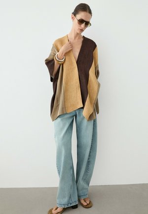 Femme portant un poncho rayé marron et beige surdimensionné, un jean bleu clair à jambes larges, des lunettes de soleil, des bracelets et des talons ouverts marron, debout contre un mur blanc.